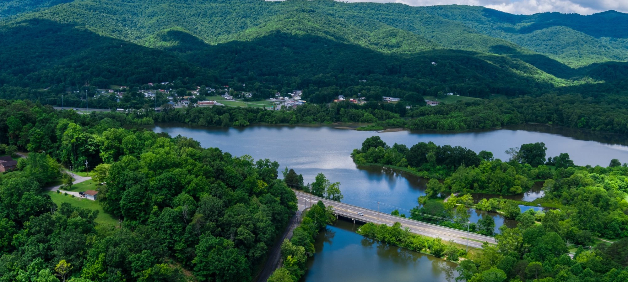 Lush green mountains, a lake, and a road bridge under a blue sky.