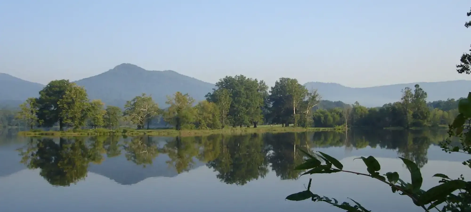 Calm lake reflecting trees and distant mountains under a clear blue sky.