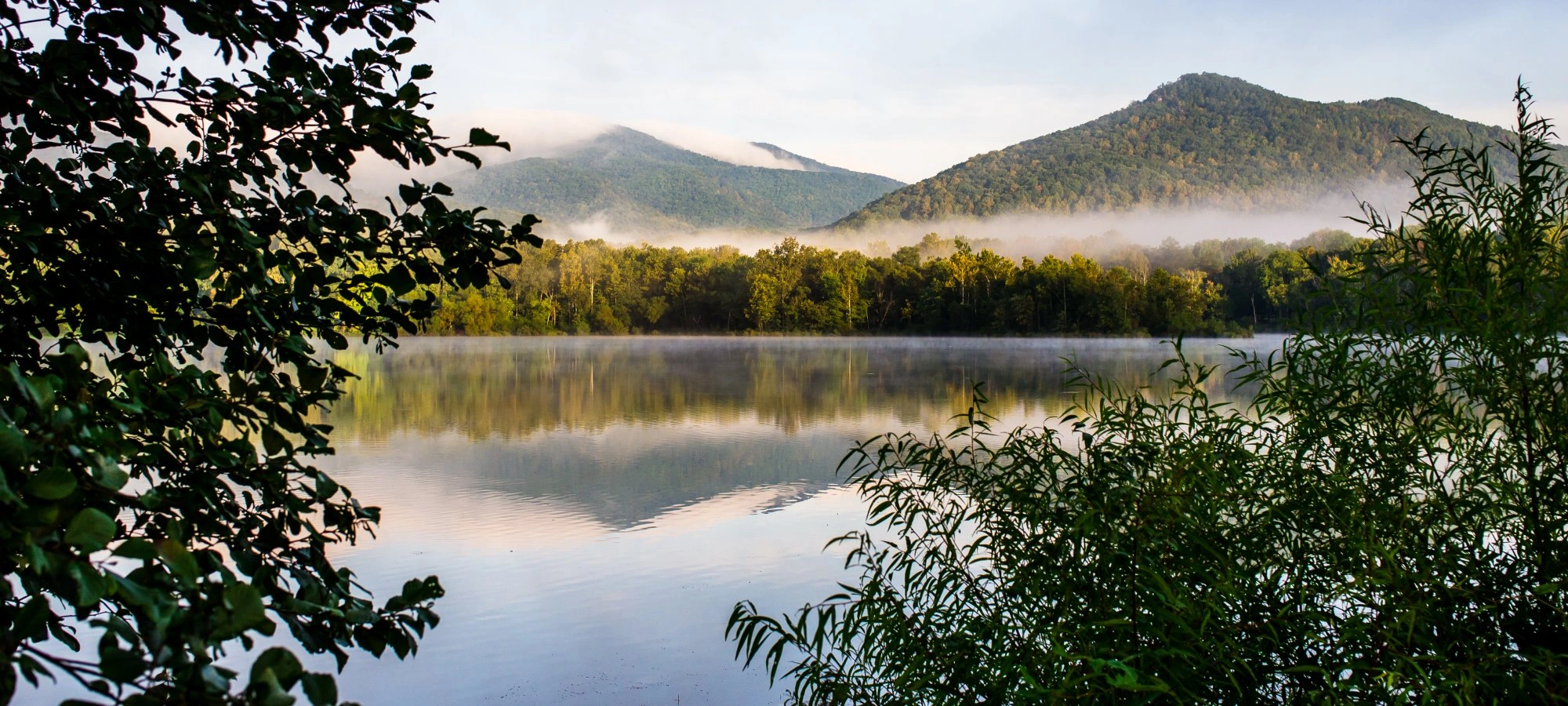 Serene lake with misty mountains and lush greenery in the foreground.