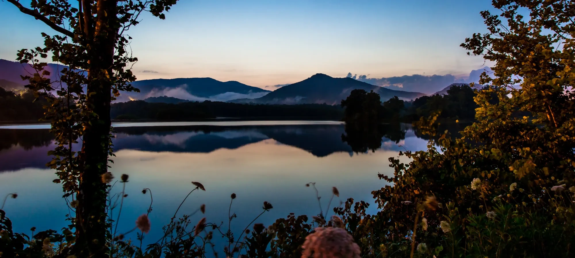 Sunset over a calm lake, mountains and trees reflected in water.