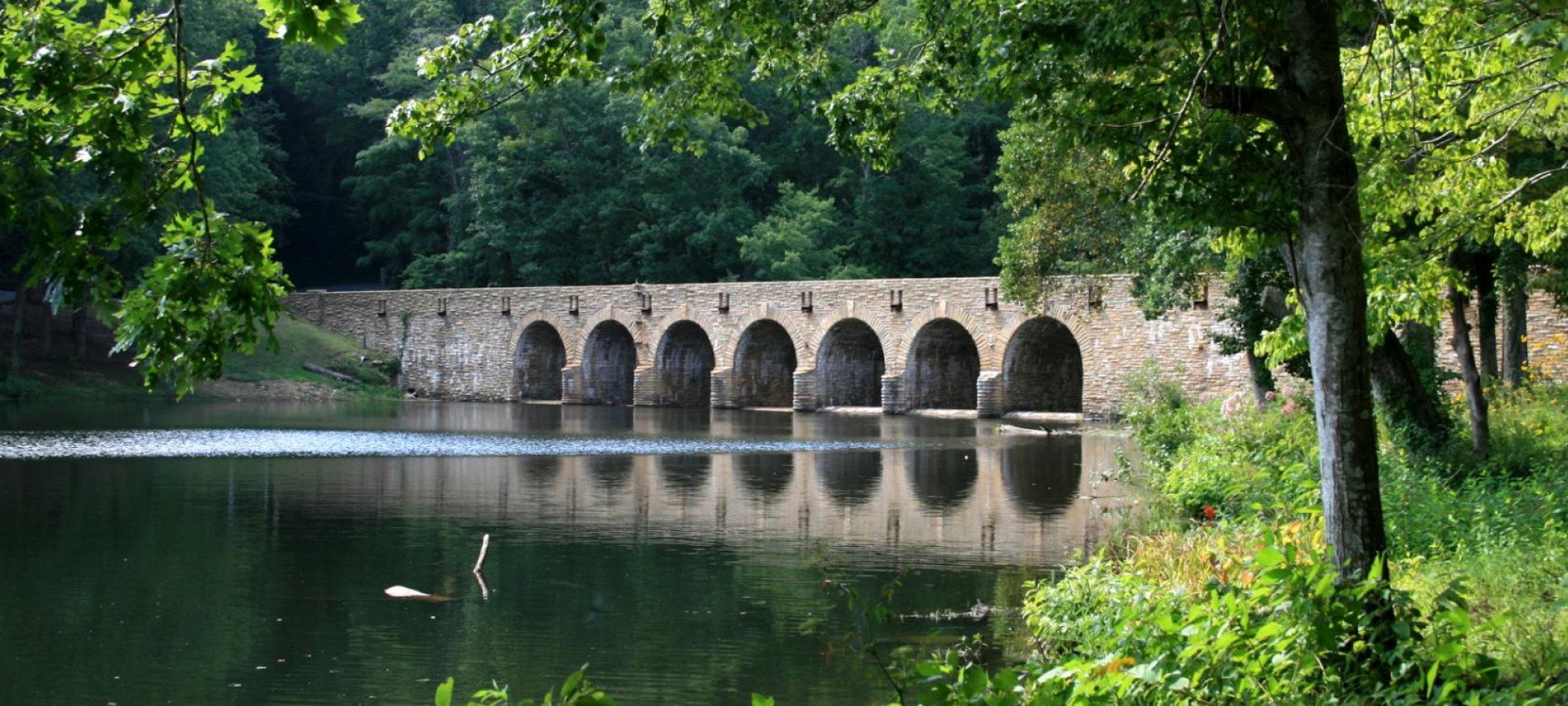 The same long arched stone bridge spans a calm lake, surrounded by dense green summer foliage.