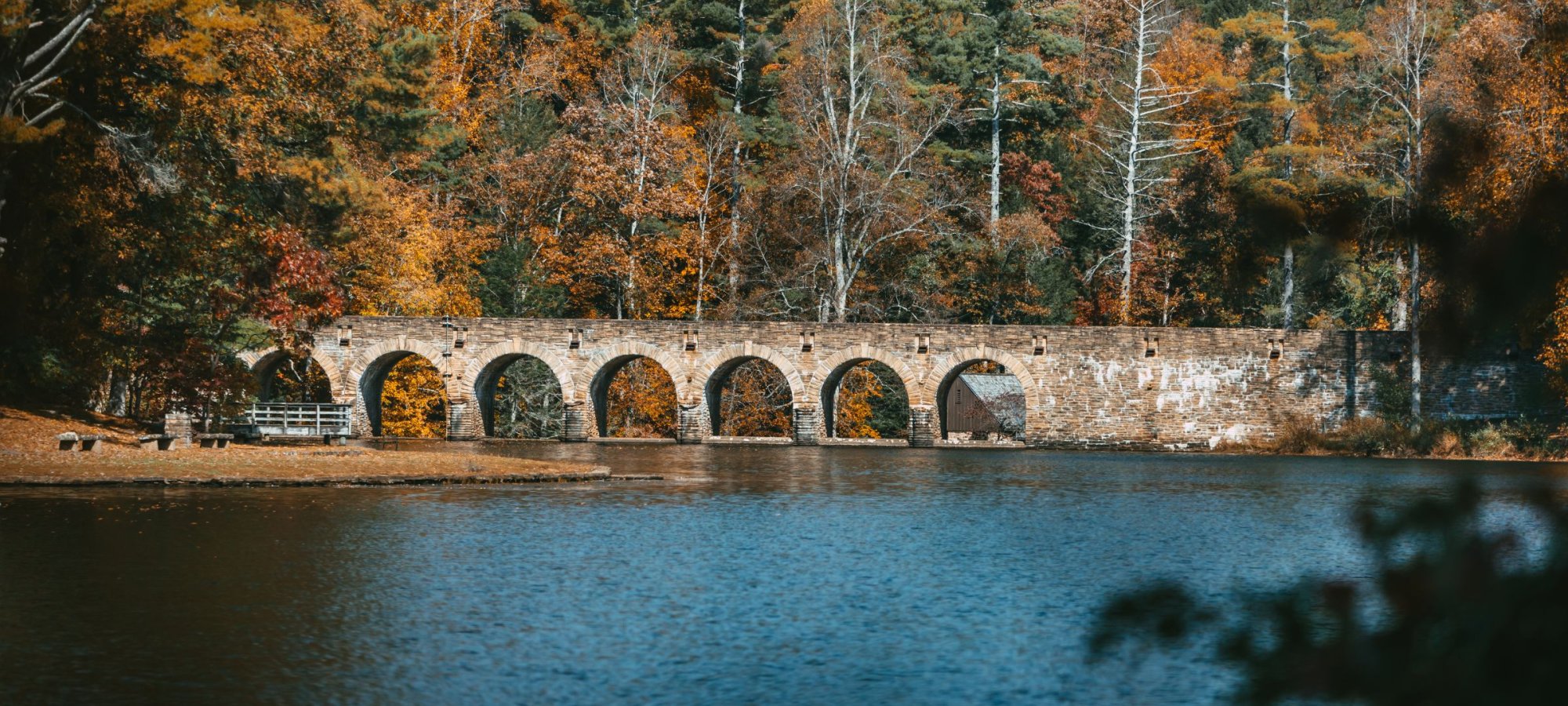 A multi-arched stone bridge spans a lake, surrounded by dense forest with vibrant fall foliage in shades of orange, yellow, and green.