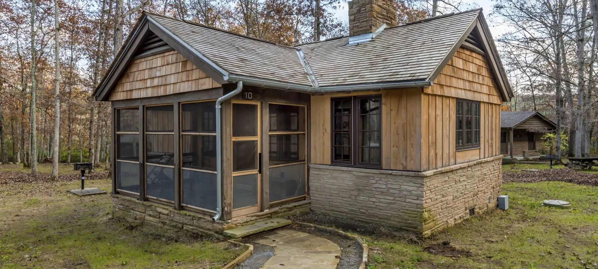 Single-story cabin with a screened porch, wood siding, and stone foundation, surrounded by bare trees in late fall.