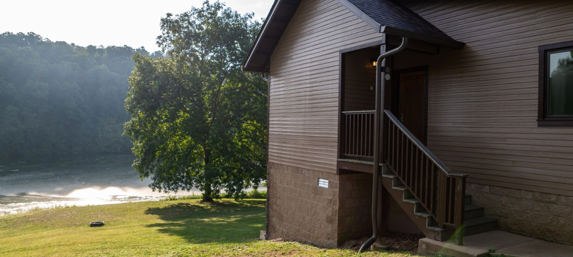 Cabin with stairs facing a tree and lake on a sunny day.