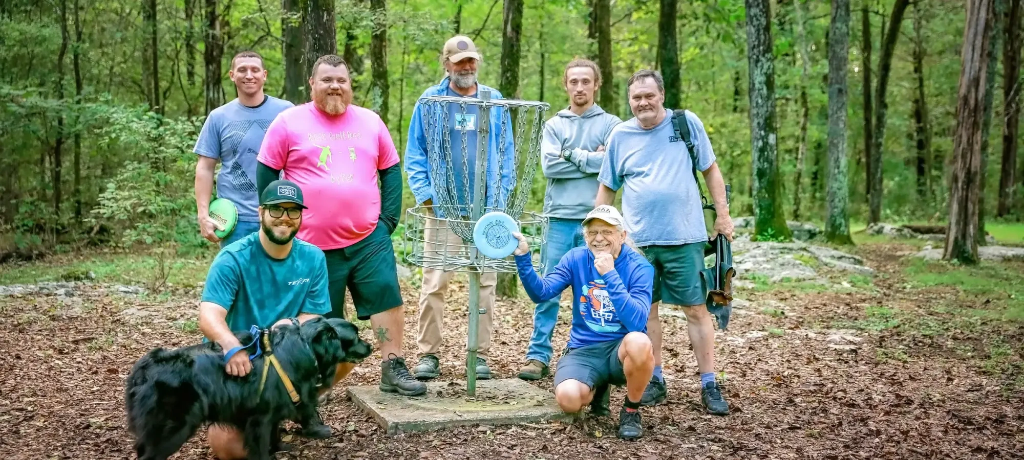 Group of men and a dog around a disc golf basket in a wooded area.