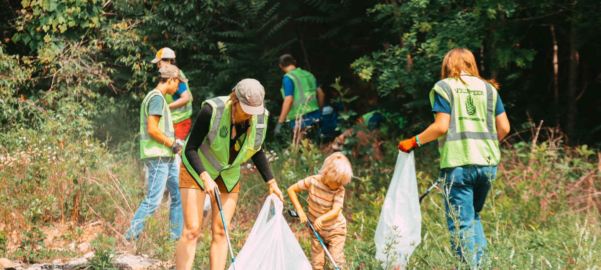 A group of volunteers, including a child, clean a wooded area. They're wearing green vests and using trash bags, conveying teamwork and environmental care.