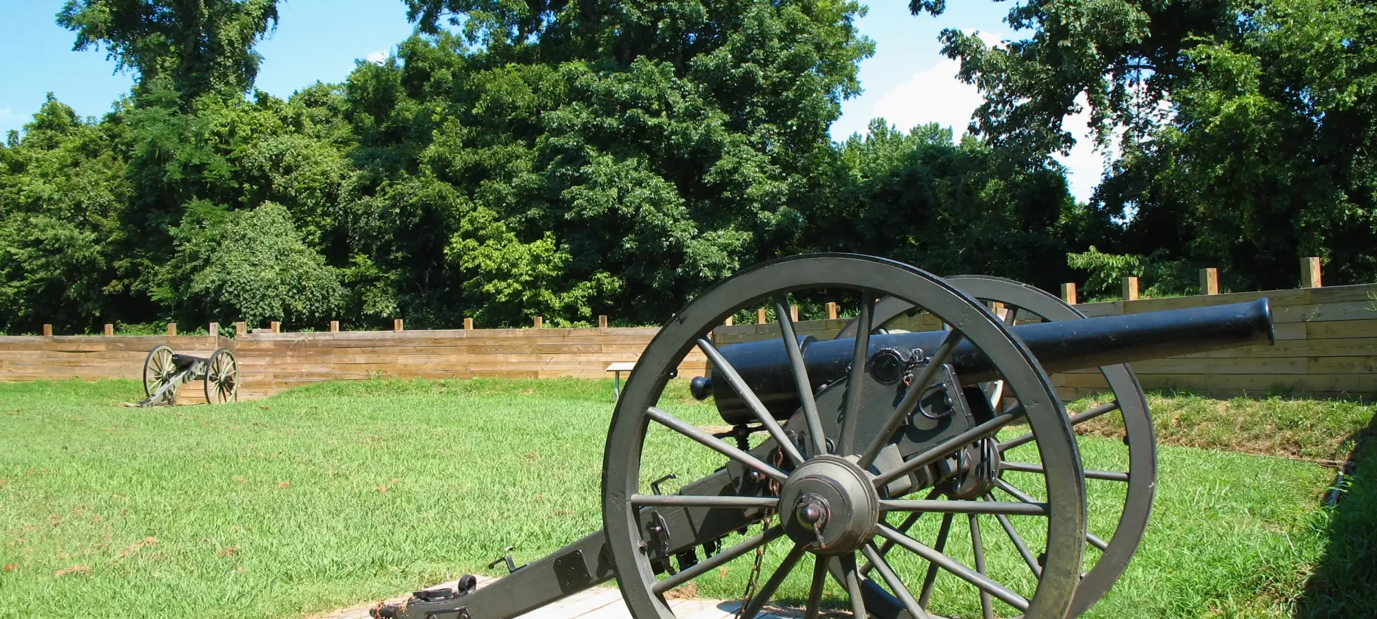 Historic cannon on grassy field with large wheels, set against a backdrop of lush green trees and a clear blue sky. The scene is calm and peaceful.
