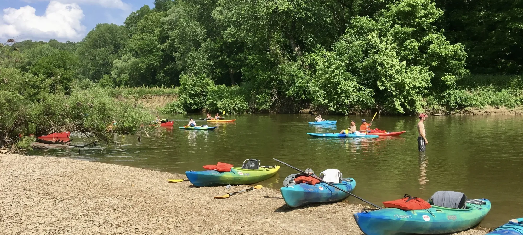 A calm river scene with several colorful kayaks on a rocky shore and others floating on the water. Green trees line the background, under a partly cloudy sky.