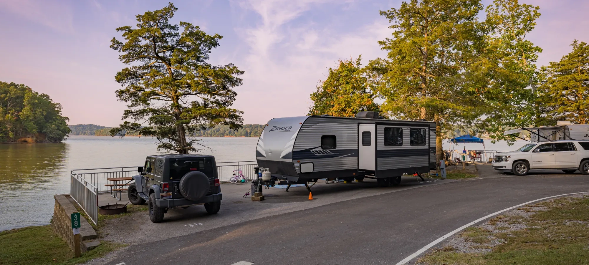 Cars and RVs parked by water and trees with a sunset in the background