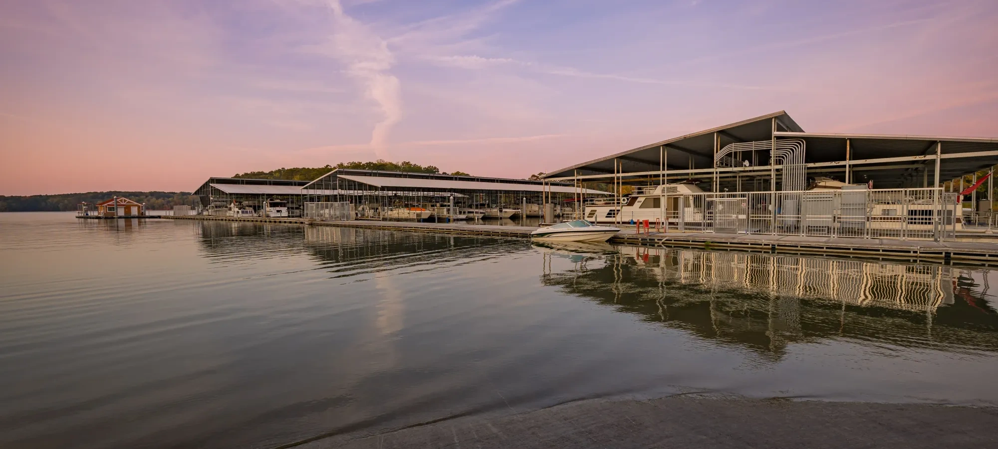 A serene marina at sunset with docks and covered boats reflected on calm water. The sky is tinged with pink and purple hues, creating a tranquil atmosphere.