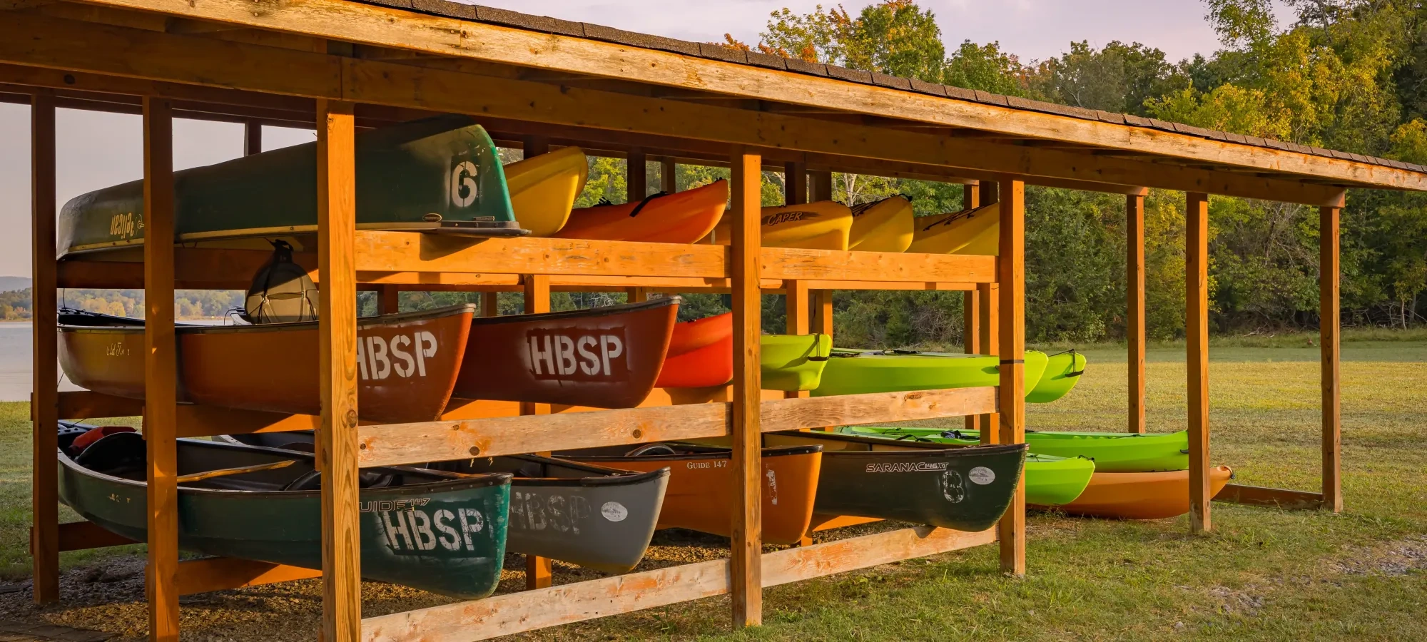 Wooden rack holding colorful kayaks, including green and orange ones, under a roof, set against a backdrop of grass and trees at sunset. Calm, outdoor scene.