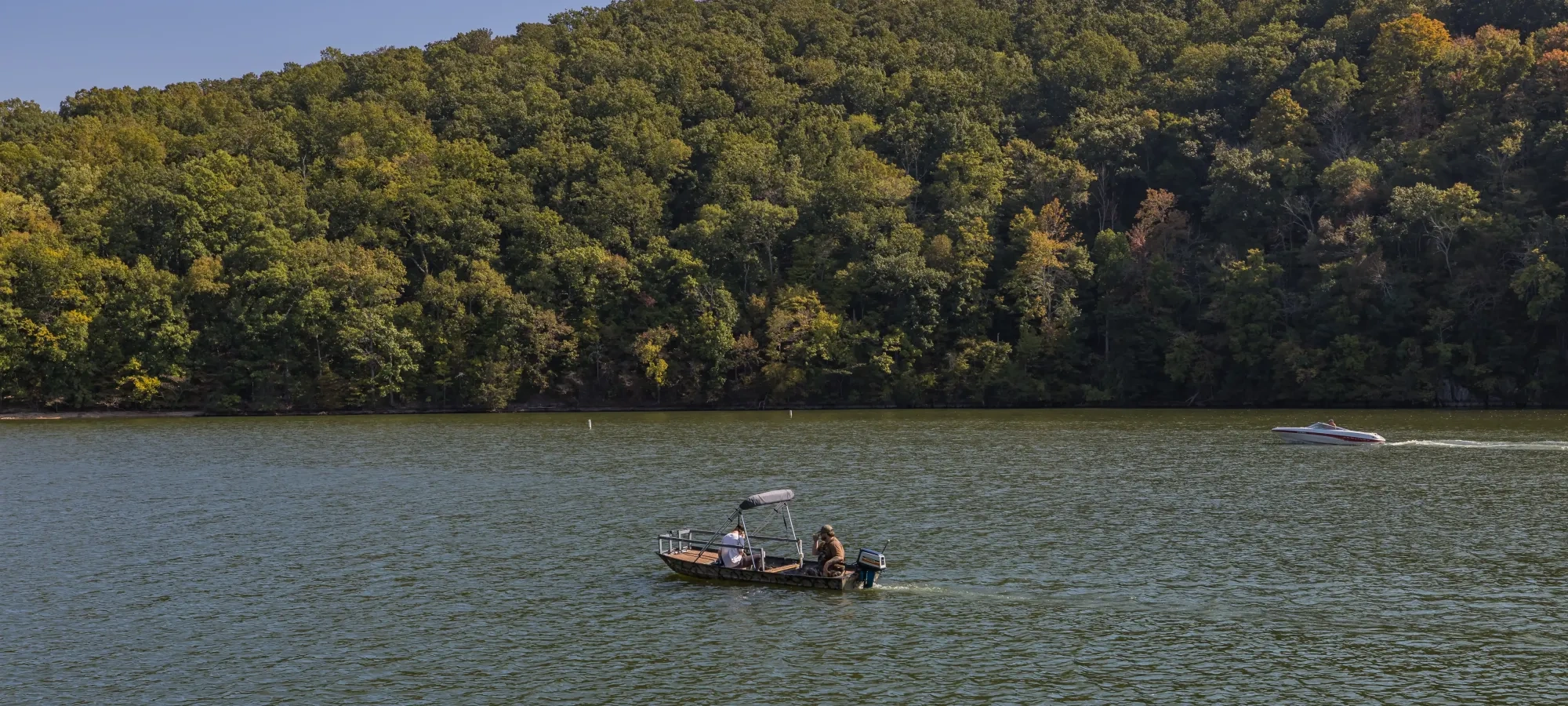Two small boats float on a peaceful lake surrounded by lush green forest under a clear blue sky. The scene conveys tranquility and natural beauty.