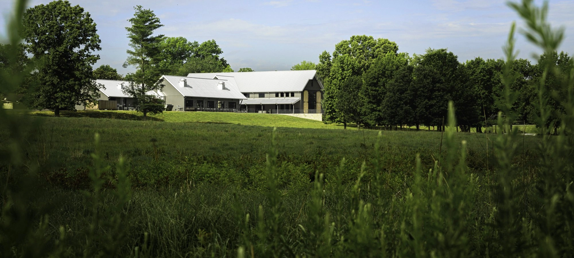 A modern lodge-style building with a metal roof surrounded by open grassy fields and trees under a blue sky.