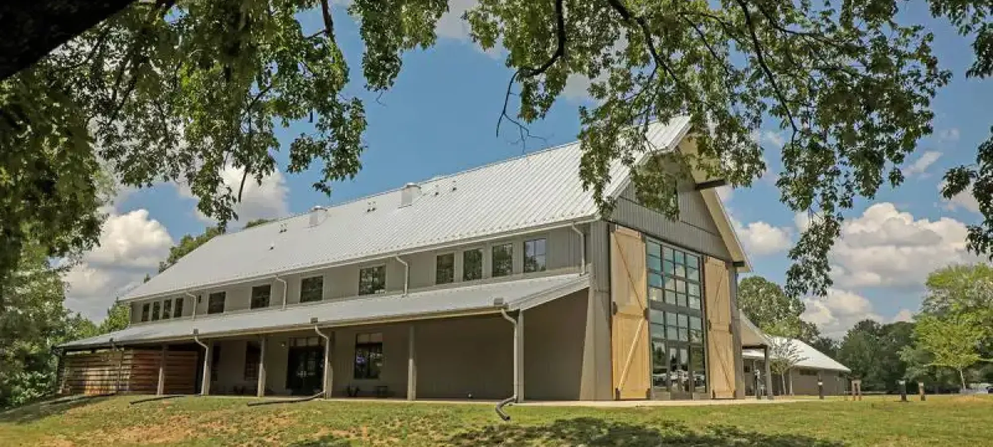 A modern barn with large glass windows and a sloped metal roof stands under a tall tree against a blue sky with clouds, evoking a serene rural atmosphere.
