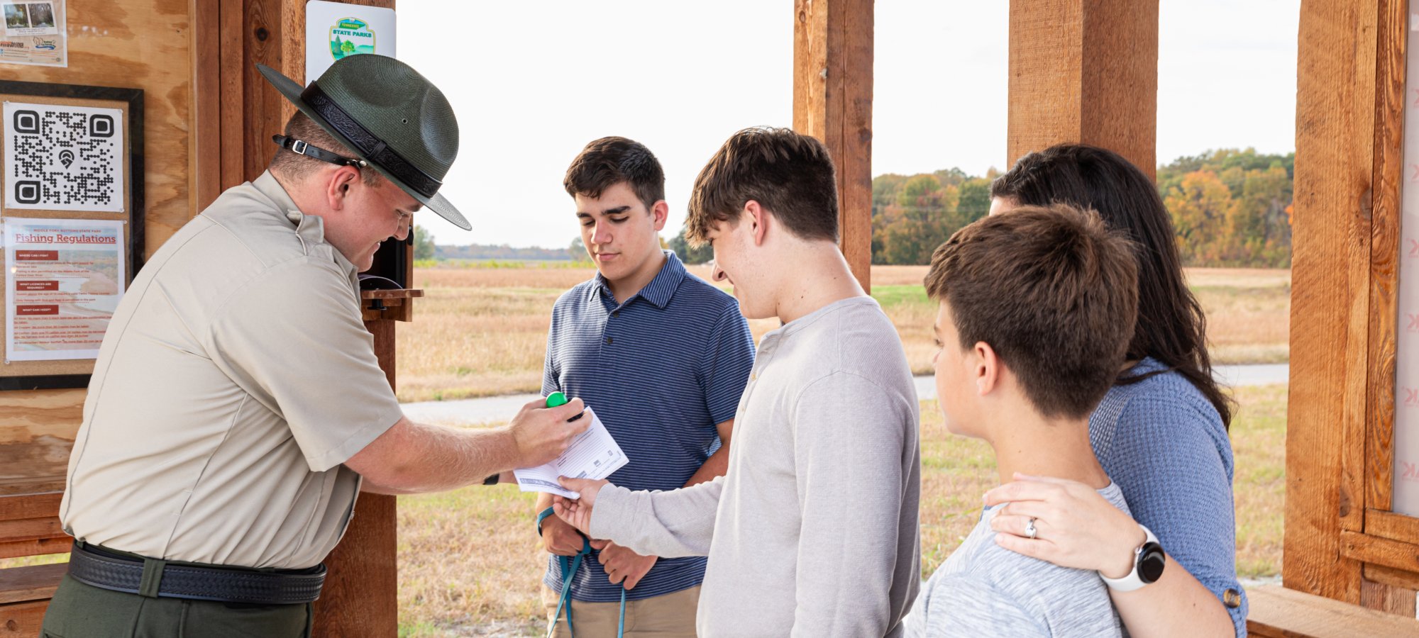 A park ranger in uniform signs a paper for four people near an open wooden shelter. The mood is friendly and relaxed, set against a rural backdrop.