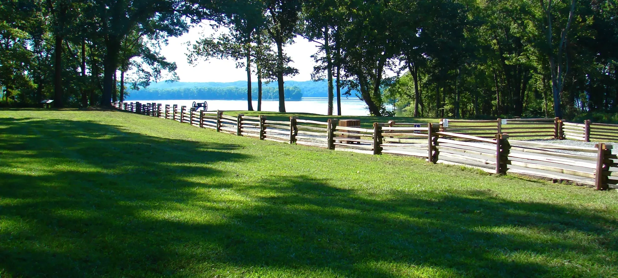 Sunny park with wooden fence, green grass, and trees by a lake.