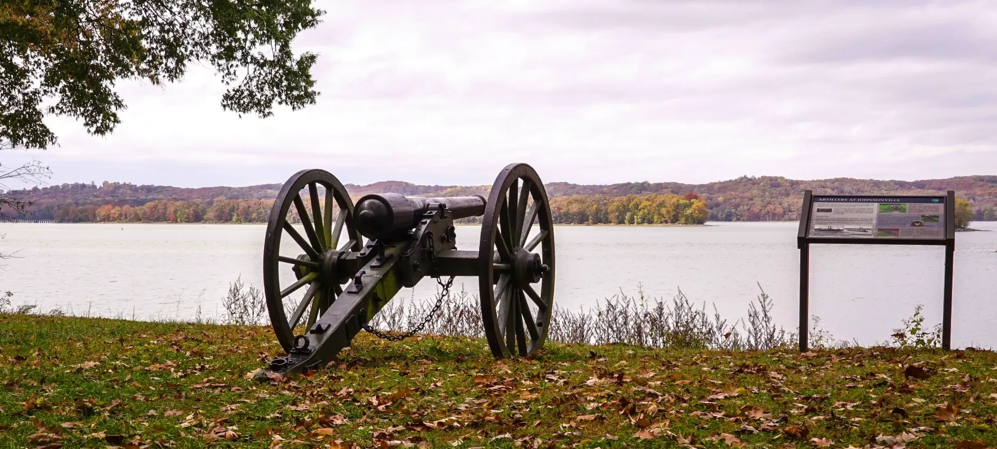 Cannon by the river with autumn leaves and hills in the background.