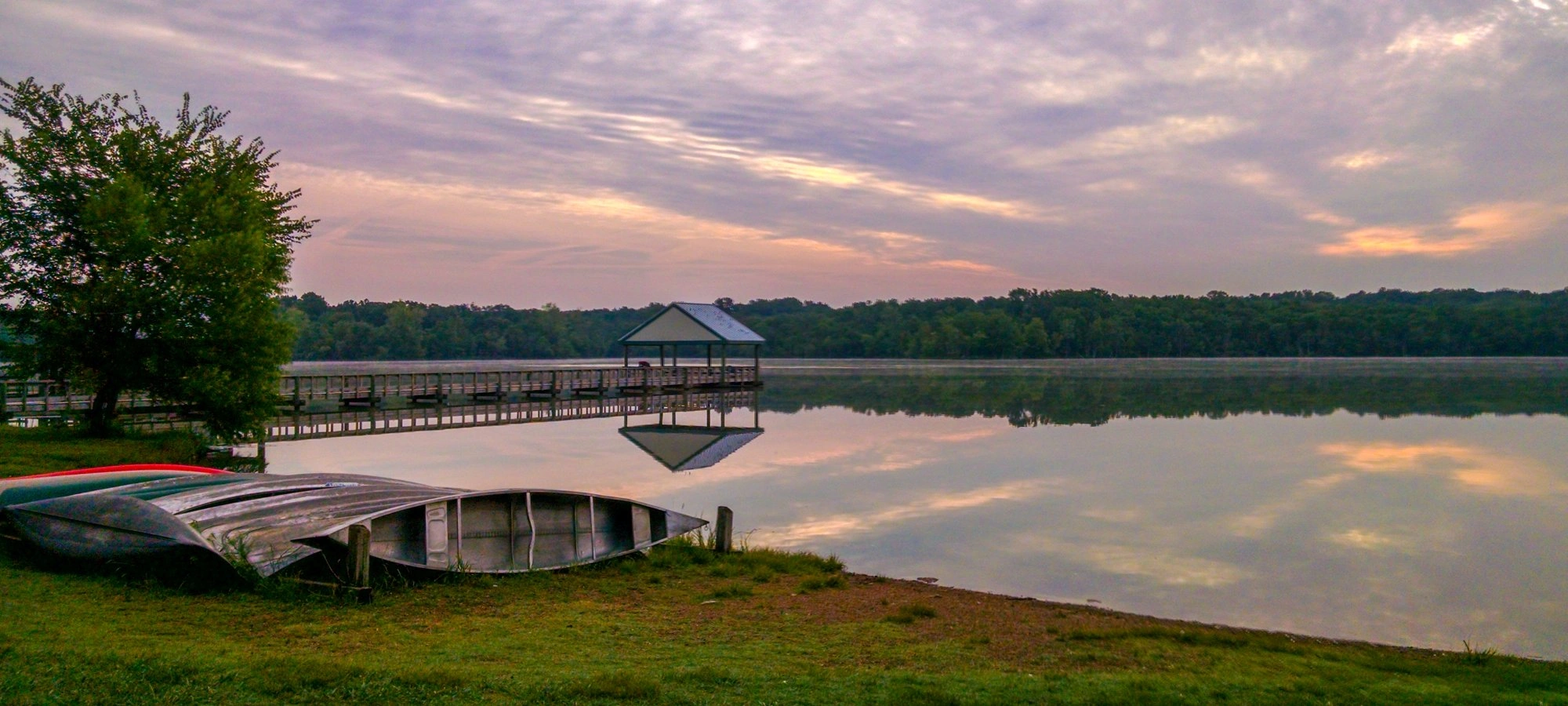 Lake at sunrise with dock, boats, and tree reflected in calm water.