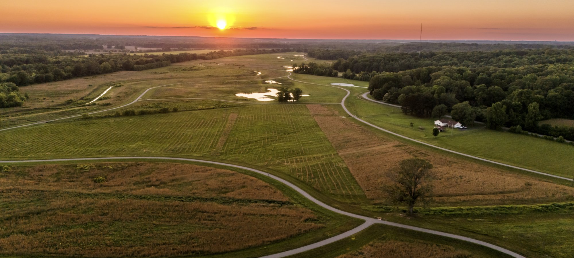 Aerial view of a serene landscape at sunset, with the sun setting over green fields, winding roads, a small pond