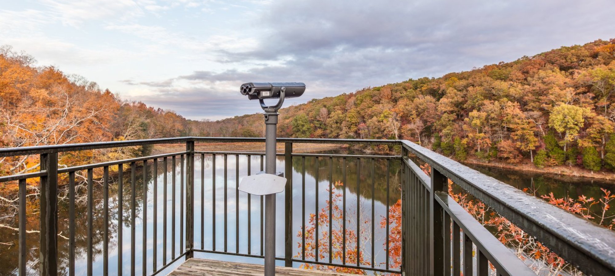 Viewing platform overlooking a calm lake surrounded by colorful autumn trees. A binocular telescope is mounted on the wooden deck