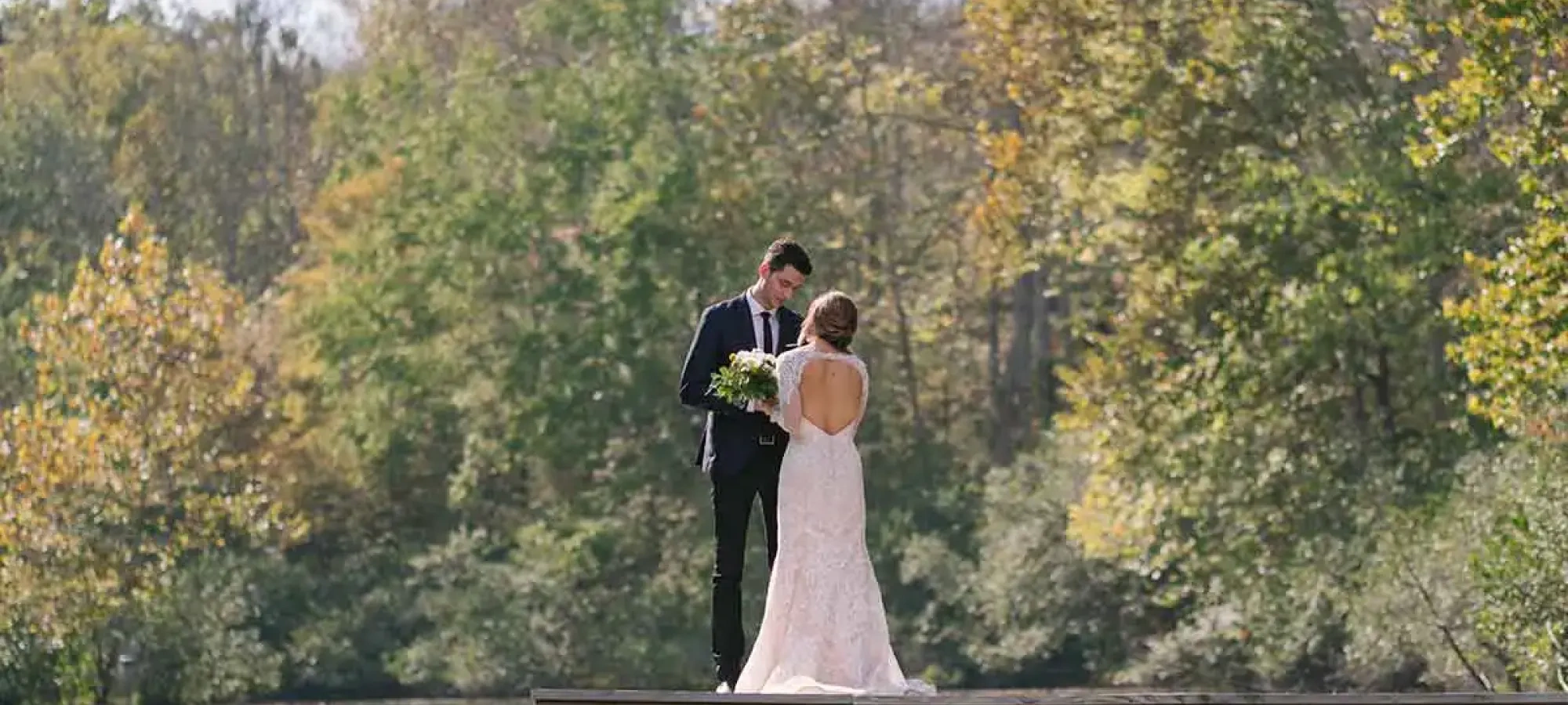 Bride and groom stand on a wooden dock, surrounded by trees and a calm lake.