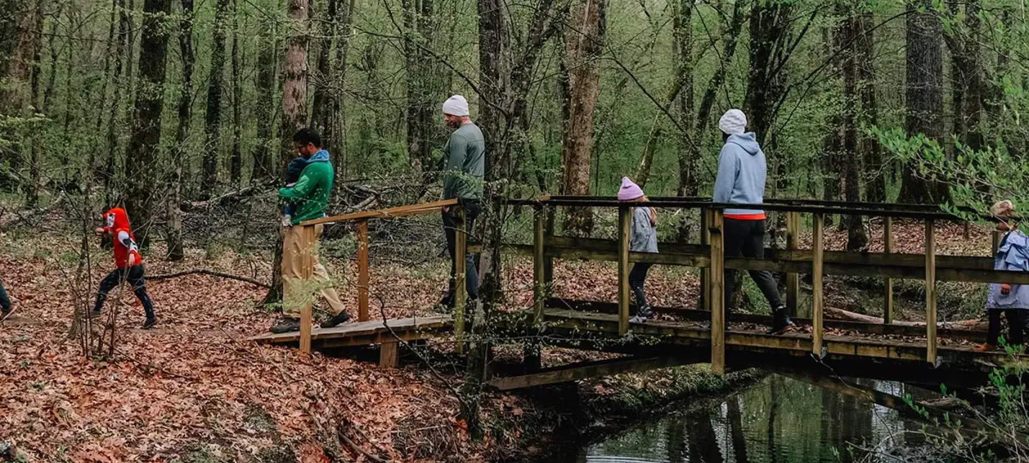 A group of people, including children and adults, walk across a small wooden bridge in a forested area with fresh green spring leaves and a shallow stream below.