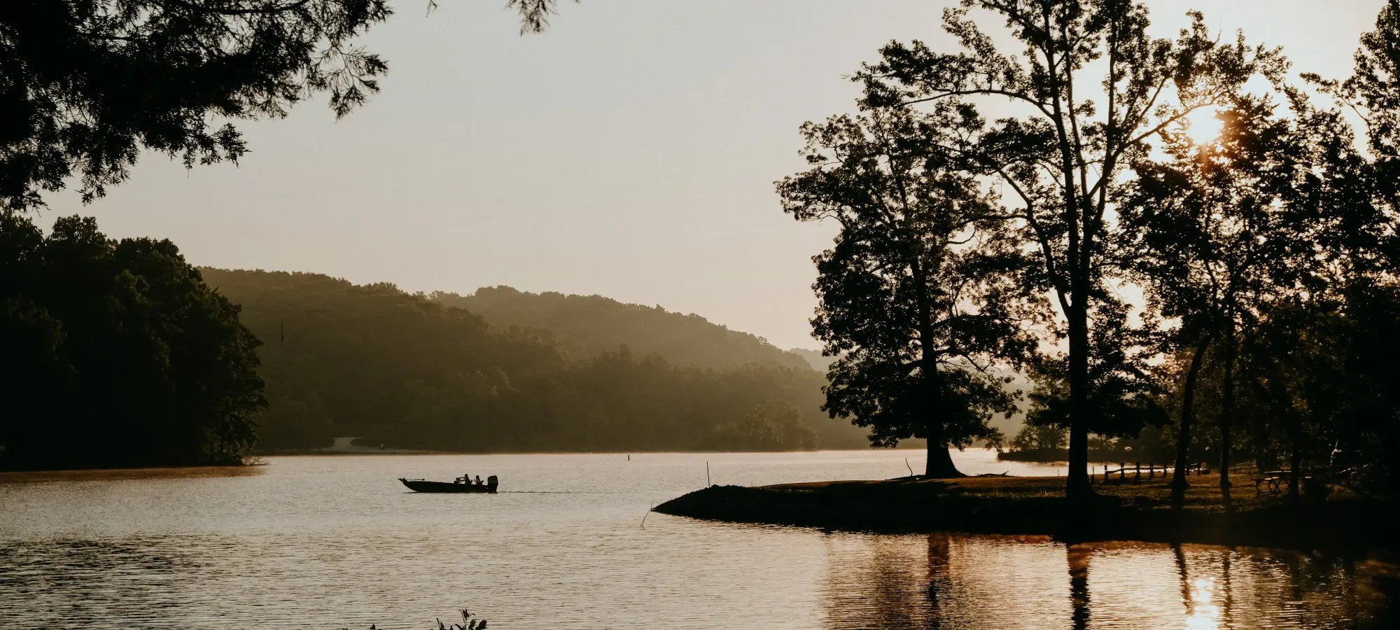 A small boat with passengers moves across a lake at sunset, with tree silhouettes and golden reflections on the water.