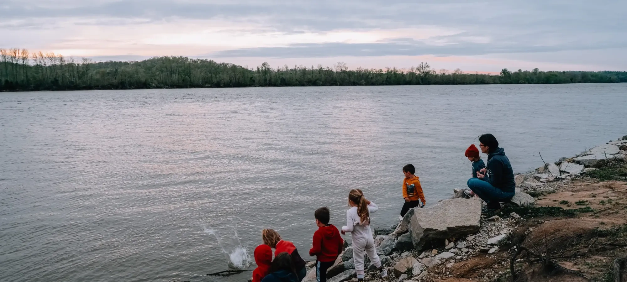 A group of children and an adult play along a rocky riverbank, tossing stones into the water under an overcast sky.