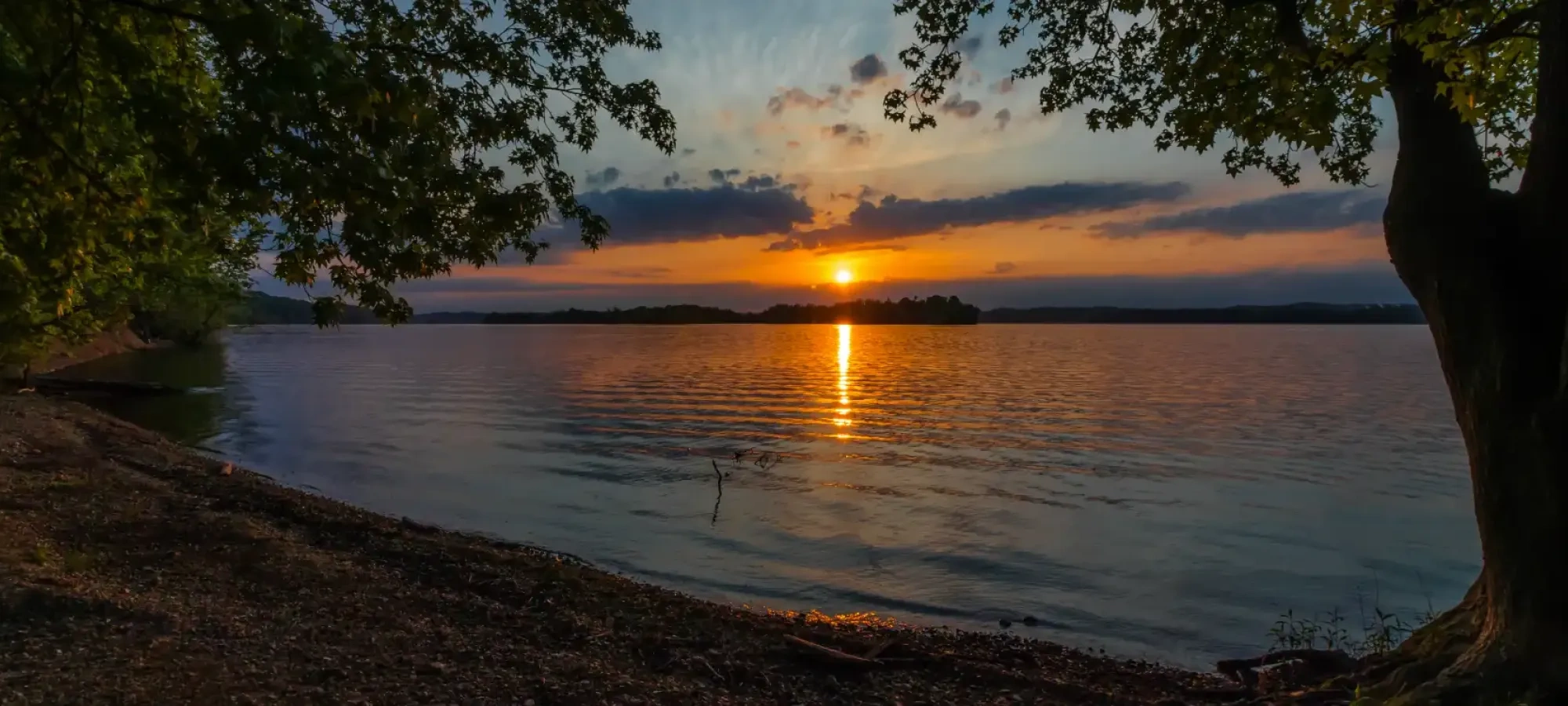 Sunset over a tranquil lake, framed by trees and a rocky shoreline.