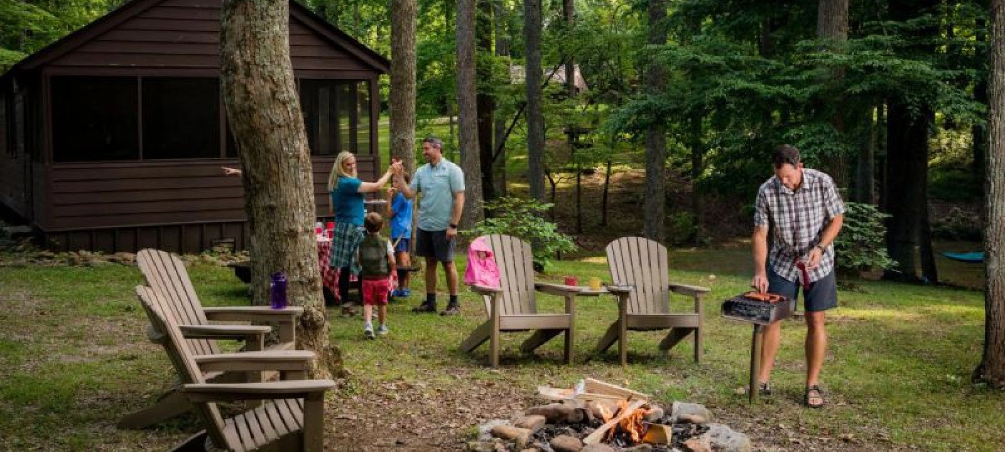 Family outside cabin in woods, man grilling by a campfire, chairs around.