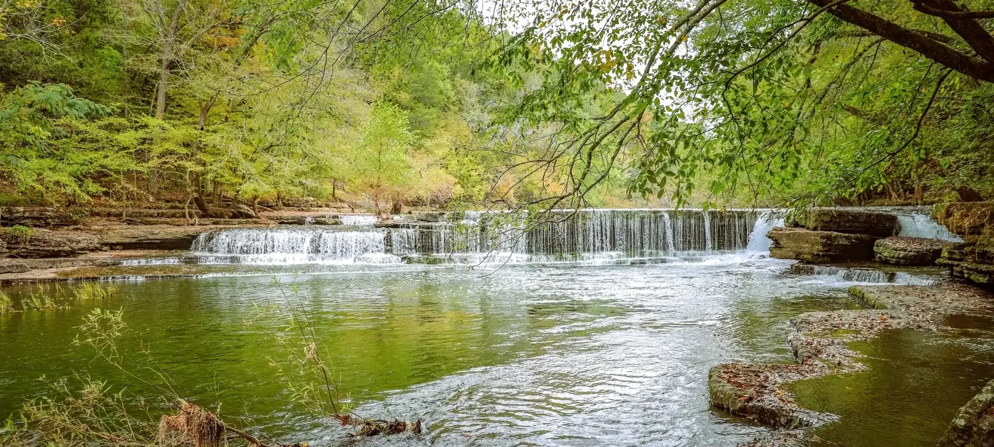A tranquil waterfall with lush green trees and calm water reflections.