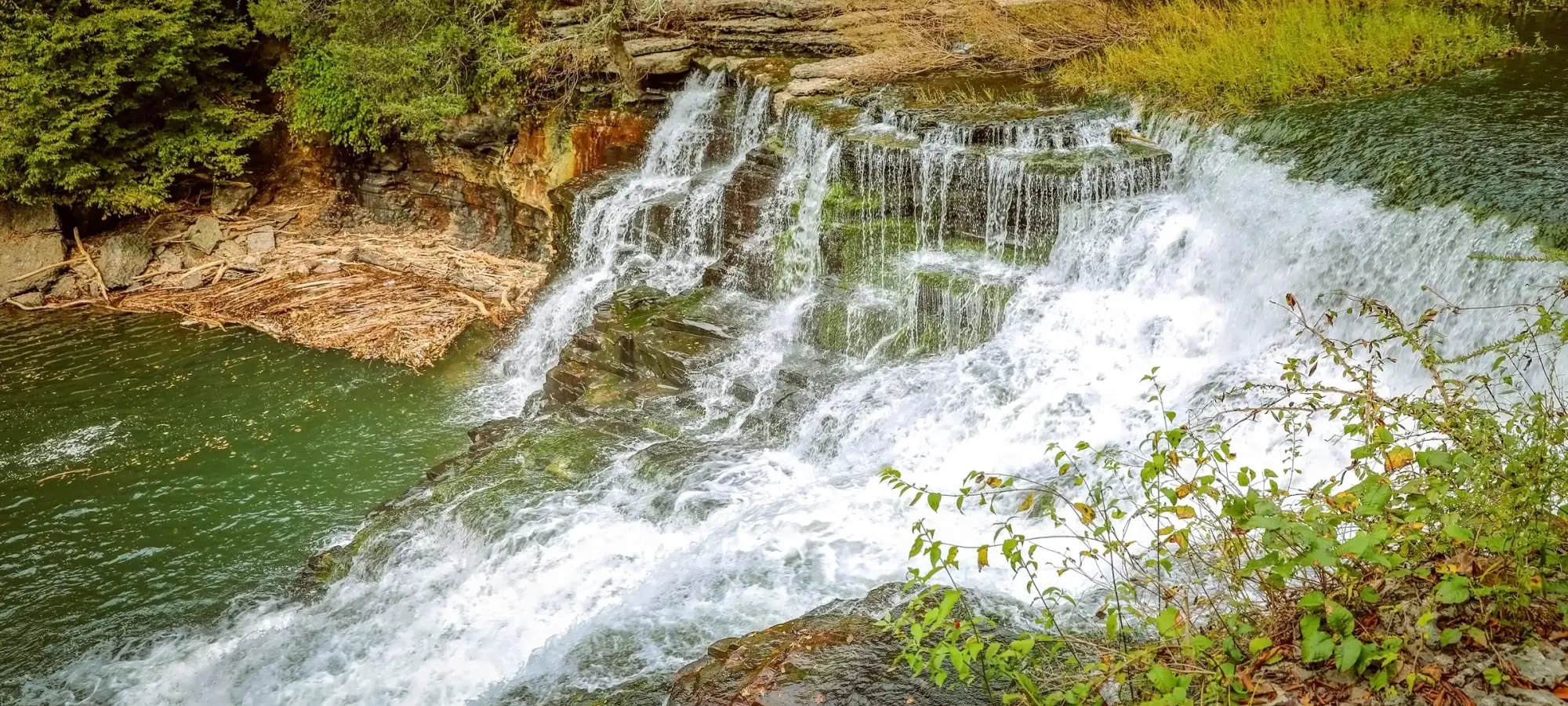 Waterfall cascading over rocks, surrounded by greenery and trees.