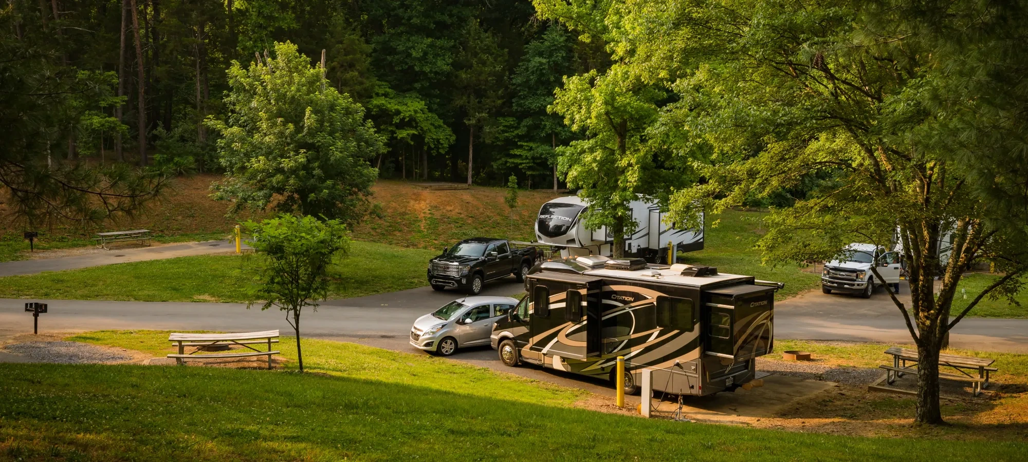 Campground scene with parked RVs and vehicles, surrounded by lush green trees.
