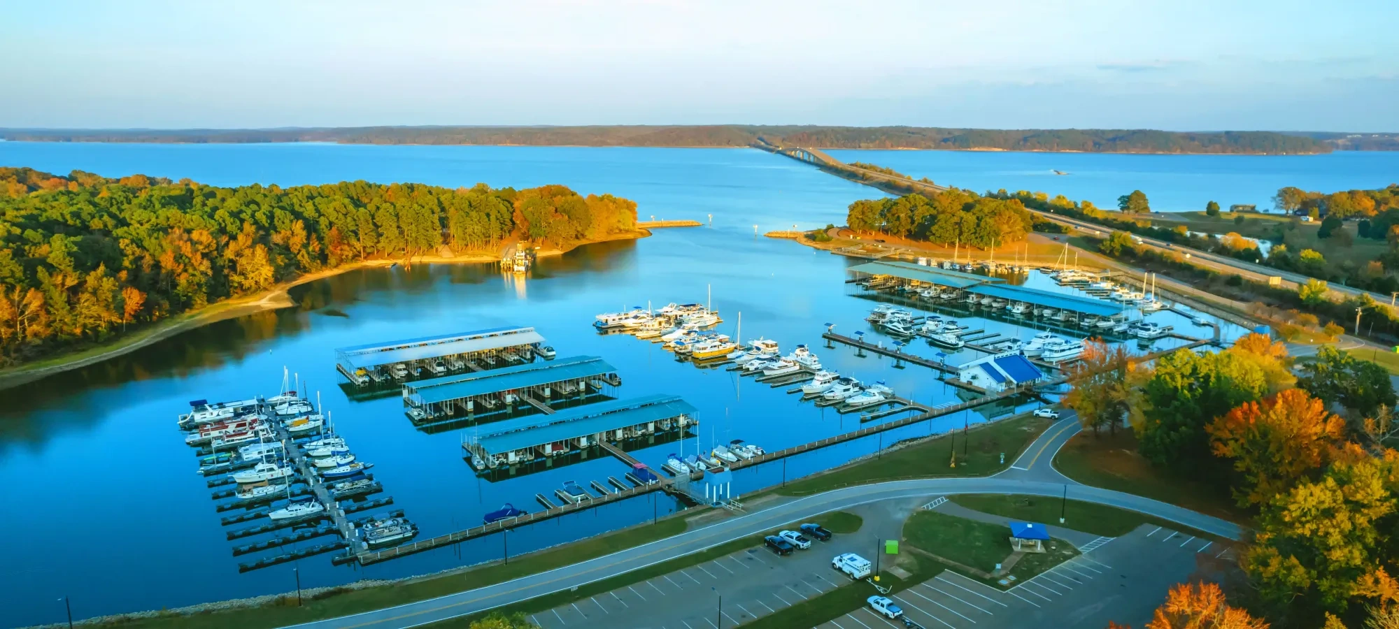 Marina with boats docked, surrounded by trees and a blue sky.