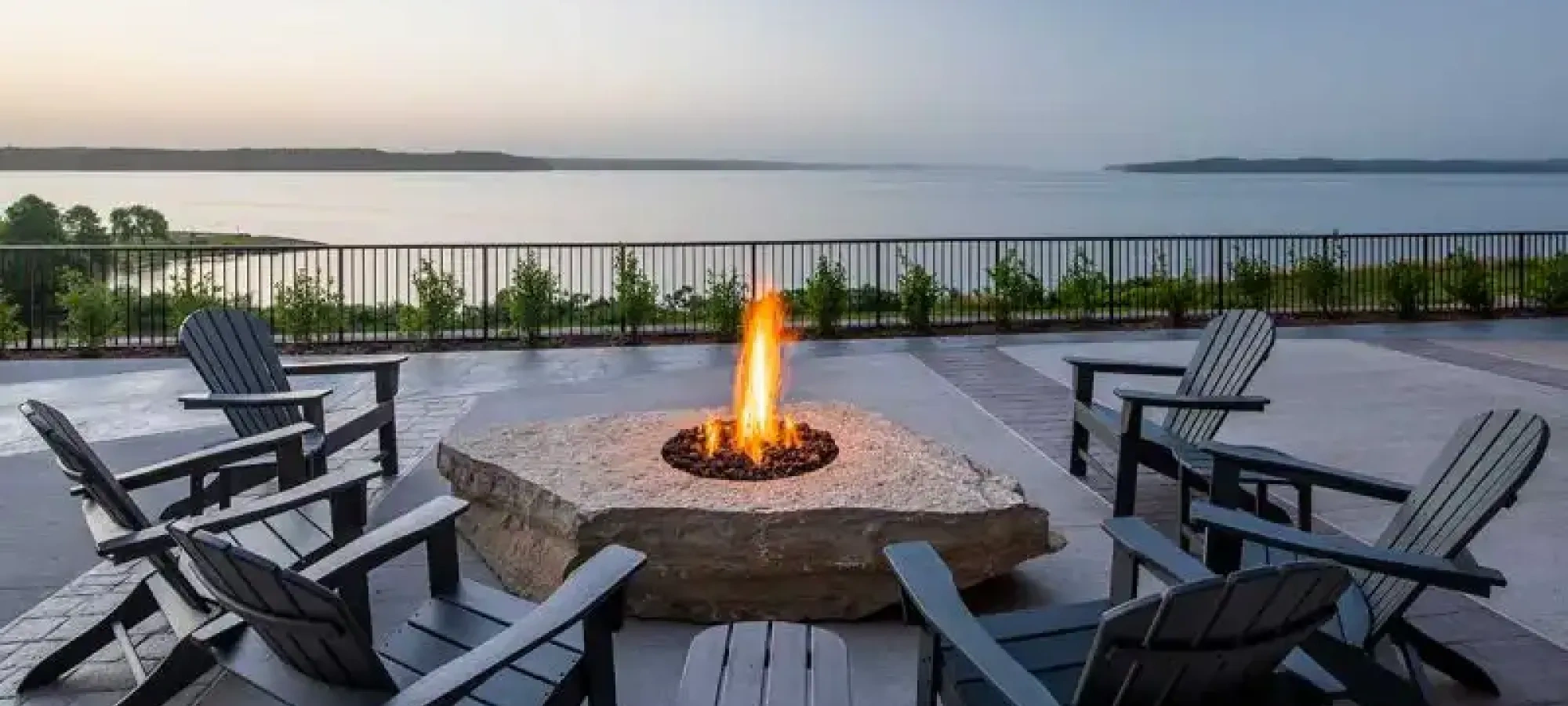Fire pit with chairs on a patio overlooking a calm lake at sunset.