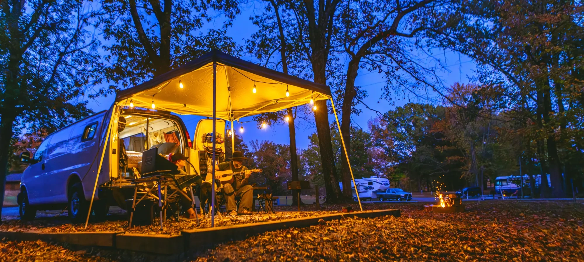 Van campsite at dusk, warm lights, people playing guitar, surrounded by autumn trees.