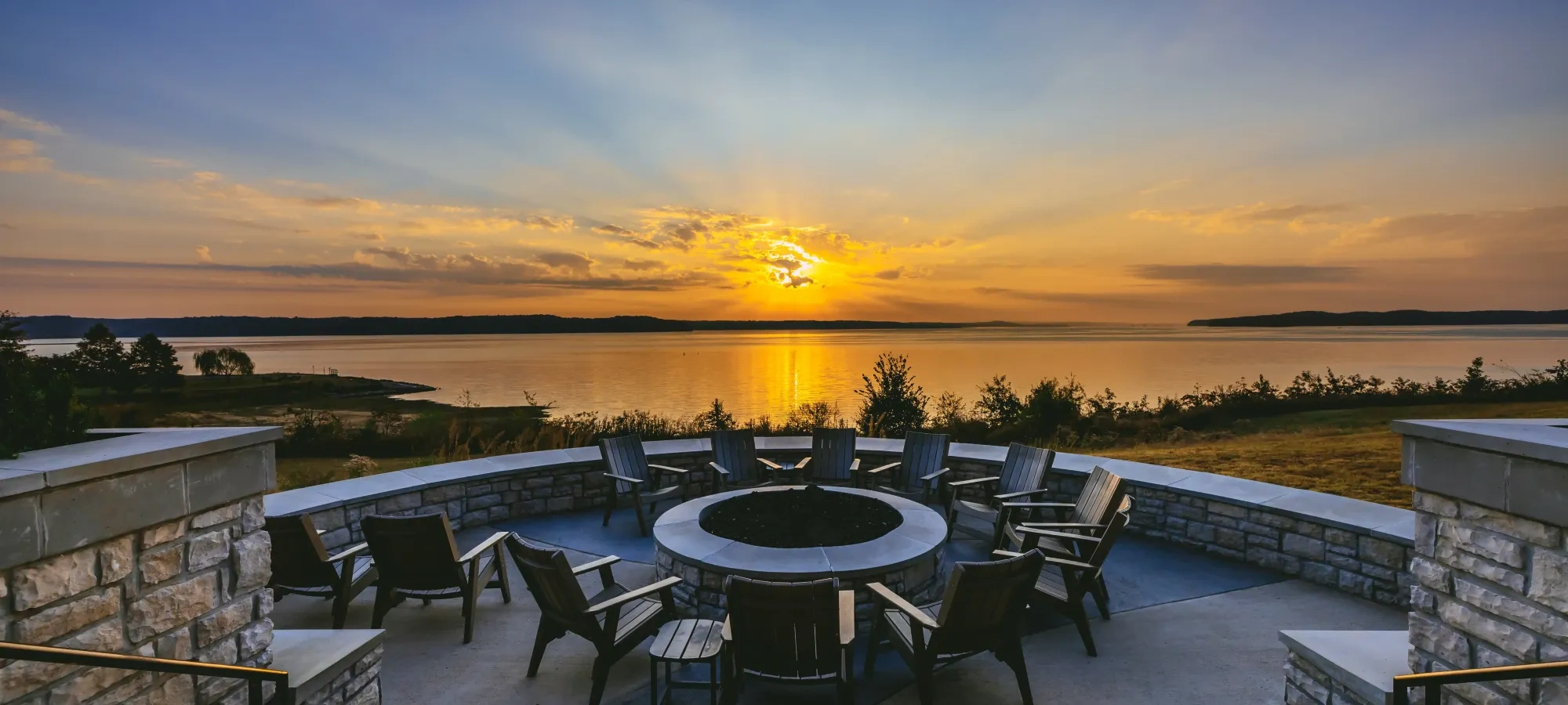 Sunset over lake with stone fire pit and chairs in foreground.