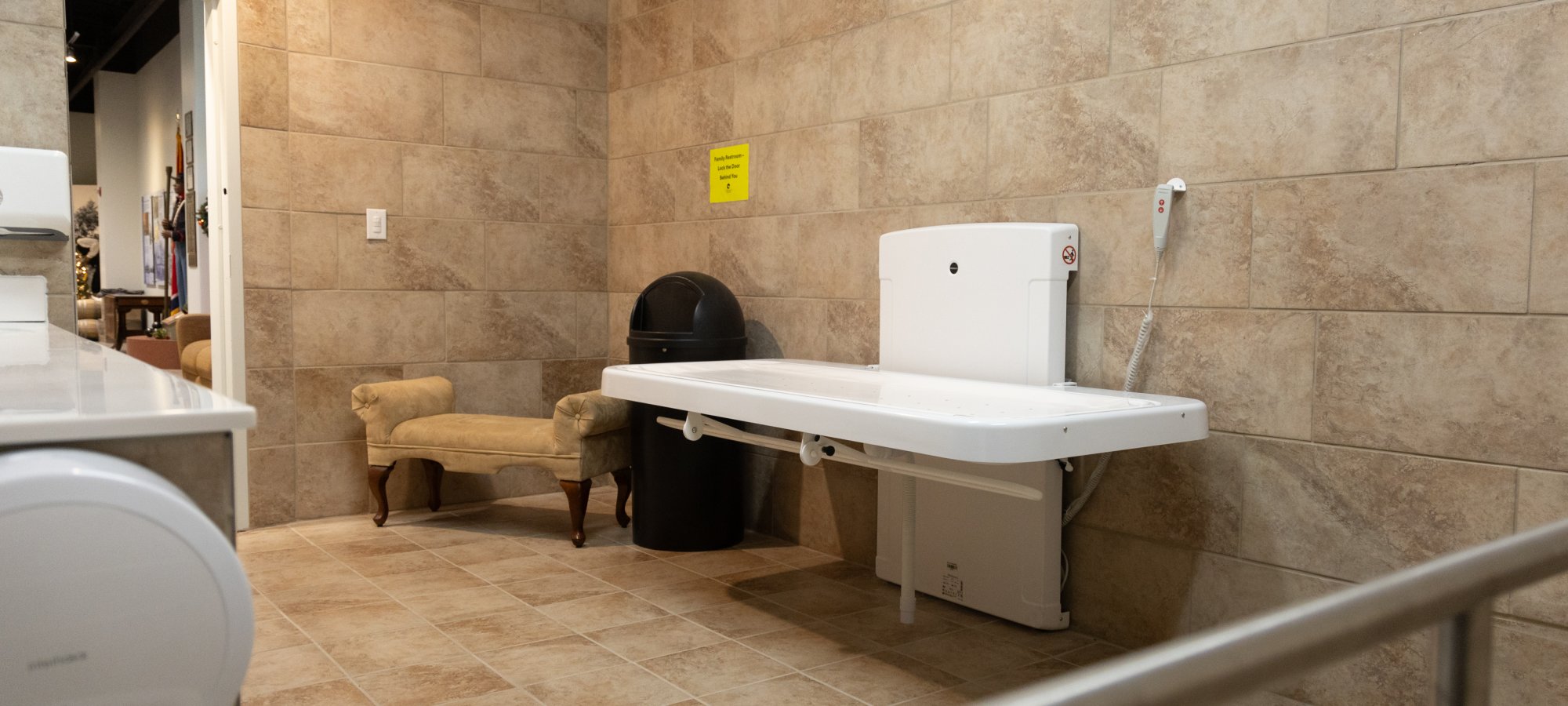 Spacious beige-tiled restroom with a wall-mounted adult changing table. A plush chair and black trash can are near the door, creating a functional, welcoming space.