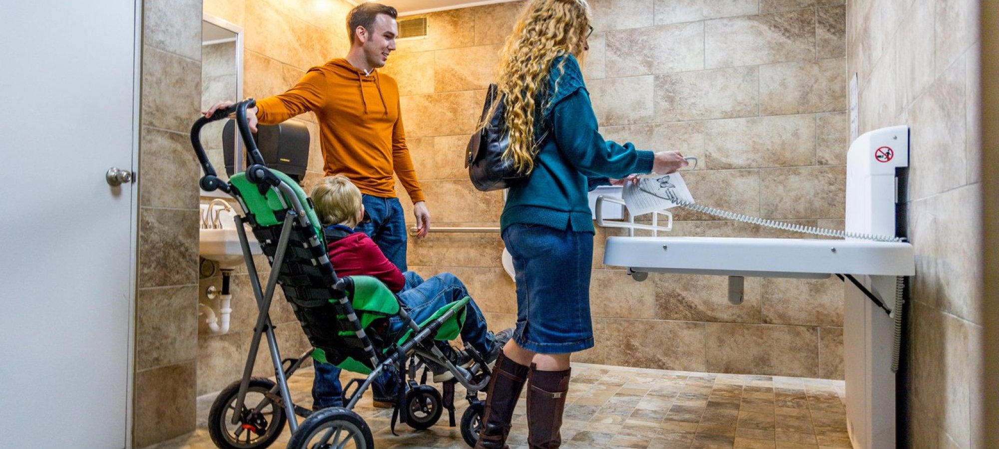 A family in a tiled bathroom with a changing table. A man holds a stroller with a child, while a woman interacts with the table. The scene is caring and relaxed.