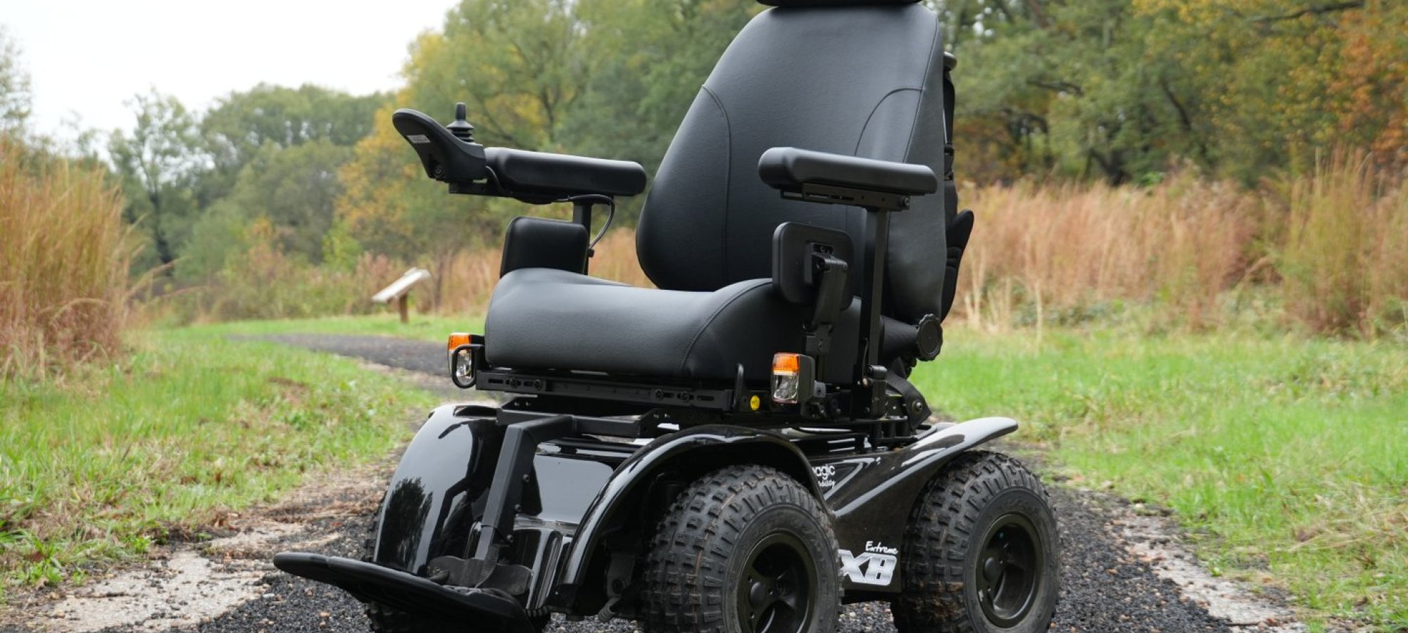 An all-terrain electric wheelchair with rugged, knobby tires on a forest path. Surrounded by grass and trees, conveying adventure and accessibility.