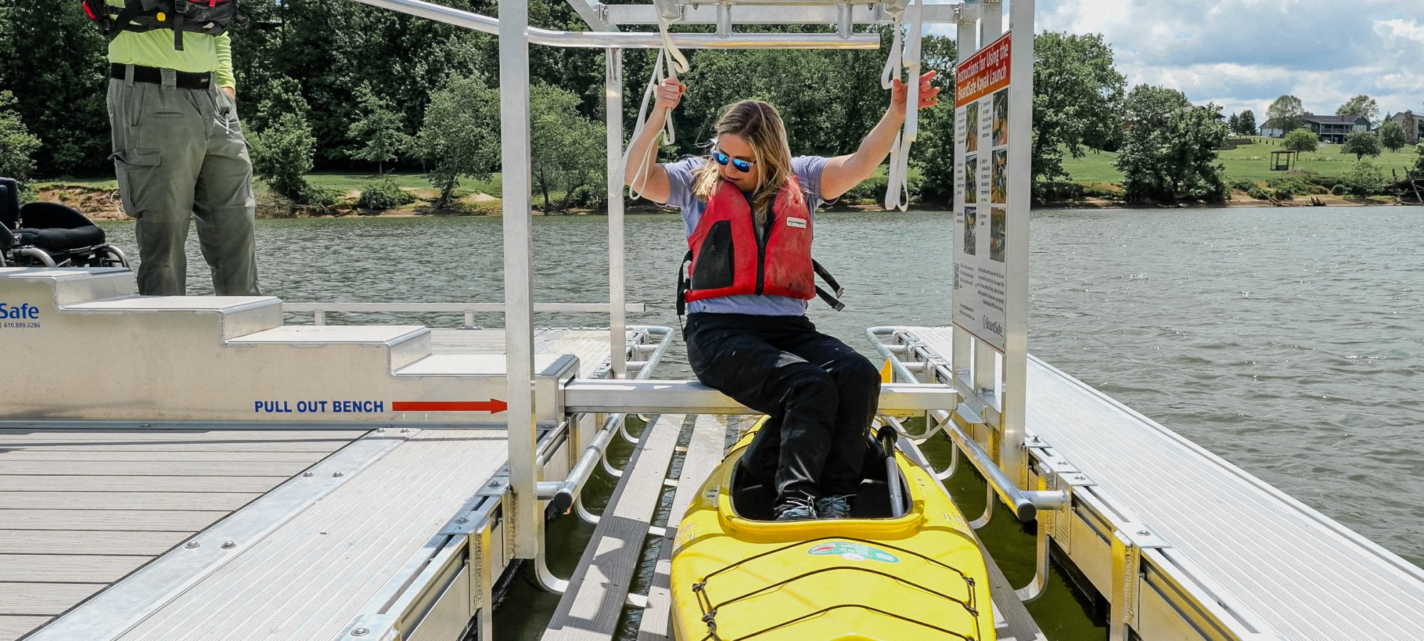 A woman in a life vest sits in a yellow kayak, using a launch mechanism on a dock by a lake. She appears focused, with green trees and cloudy skies in the background.