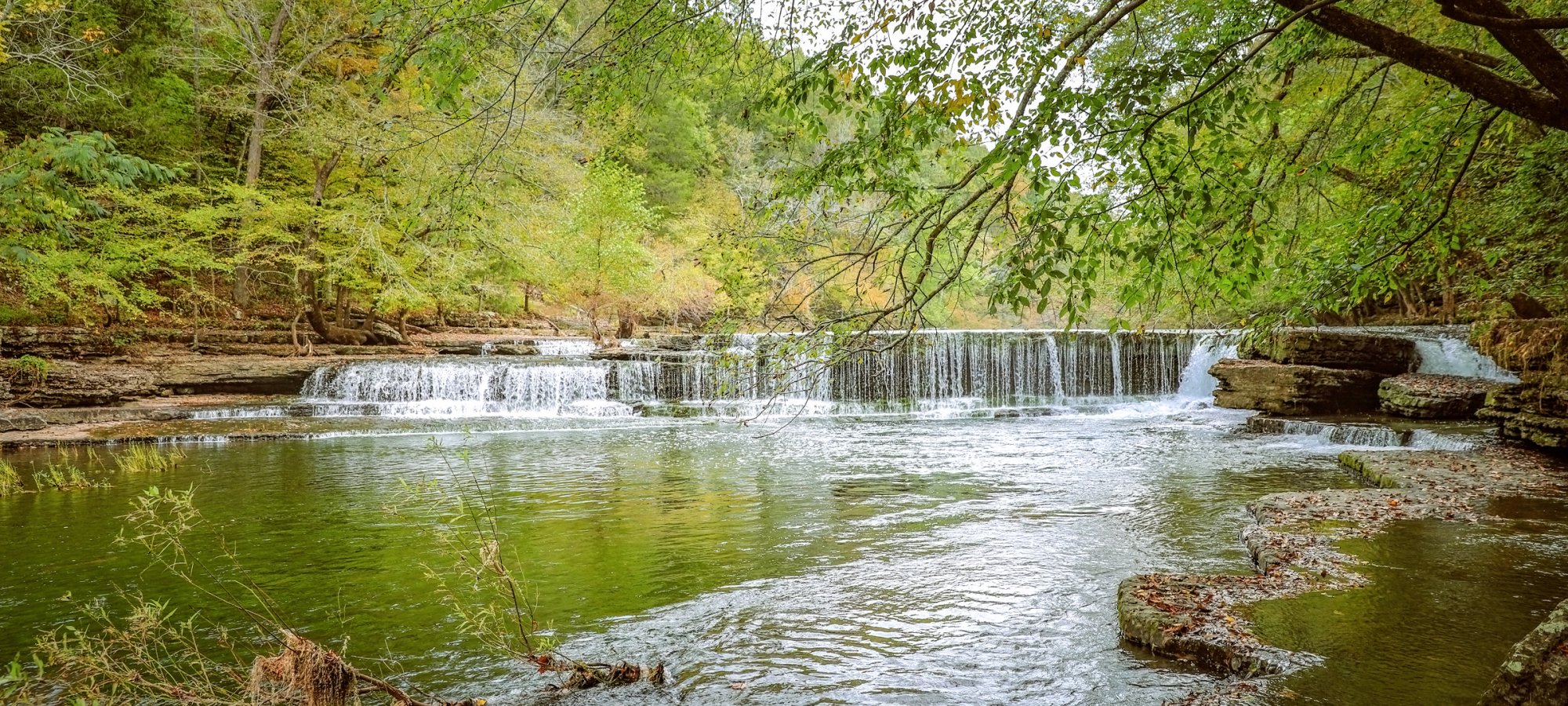 Tranquil scene of a small waterfall flowing into a clear river surrounded by lush, green trees. The calm water reflects the vibrant greenery above.