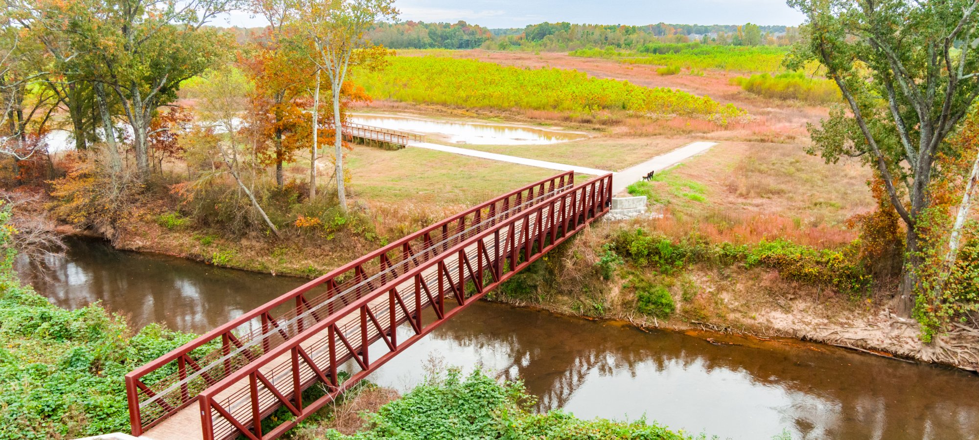 A red pedestrian bridge spans a calm river, surrounded by lush trees with autumn foliage and expansive, colorful fields under a cloudy sky.