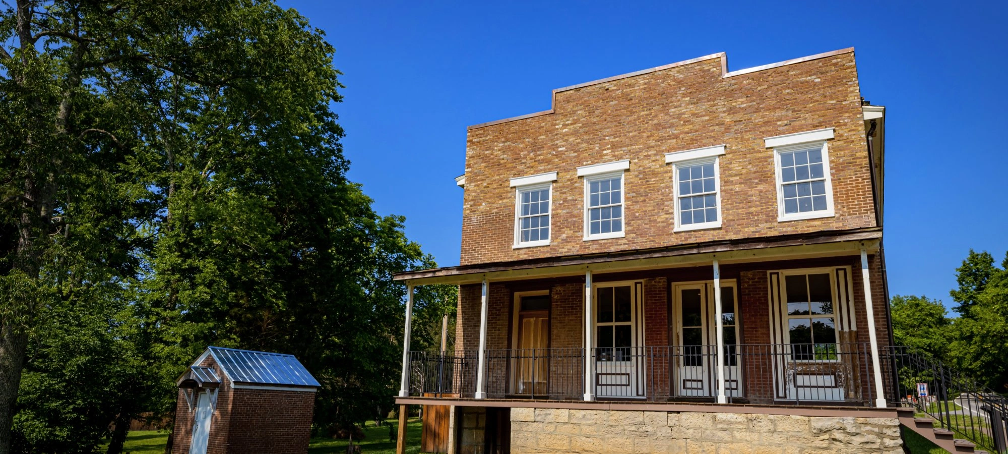 Historic brick building with large windows, surrounded by lush trees under a clear blue sky.