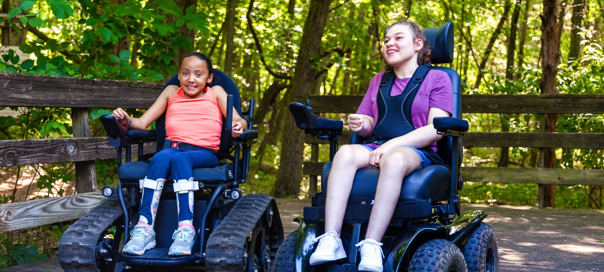 Two smiling kids in wheelchairs on a wooden path in a lush green forest.