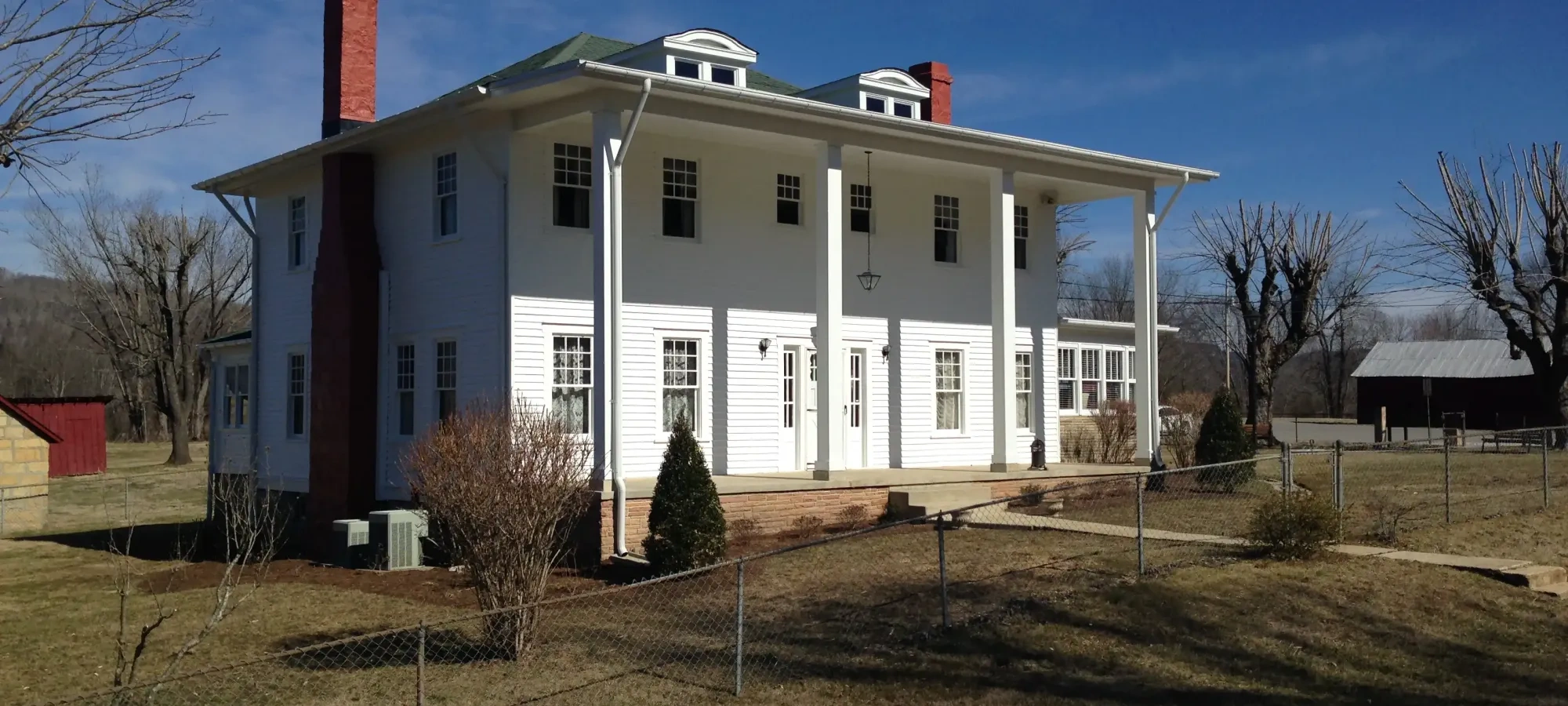 A large white colonial-style house with red brick chimneys stands under a bright blue sky, surrounded by a fenced grassy yard and bare trees.