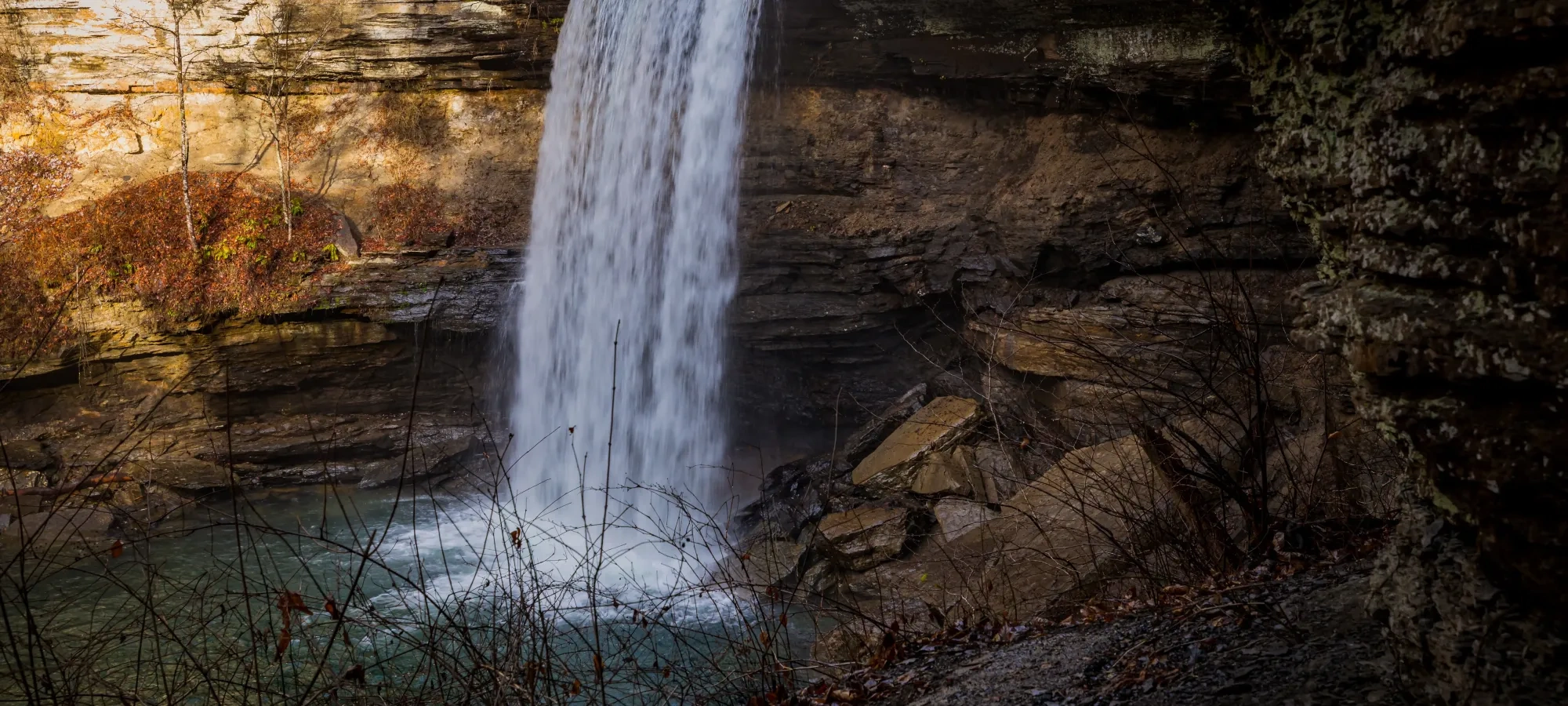 Waterfall cascading over rocky cliff into a turquoise pool, surrounded by dry branches.