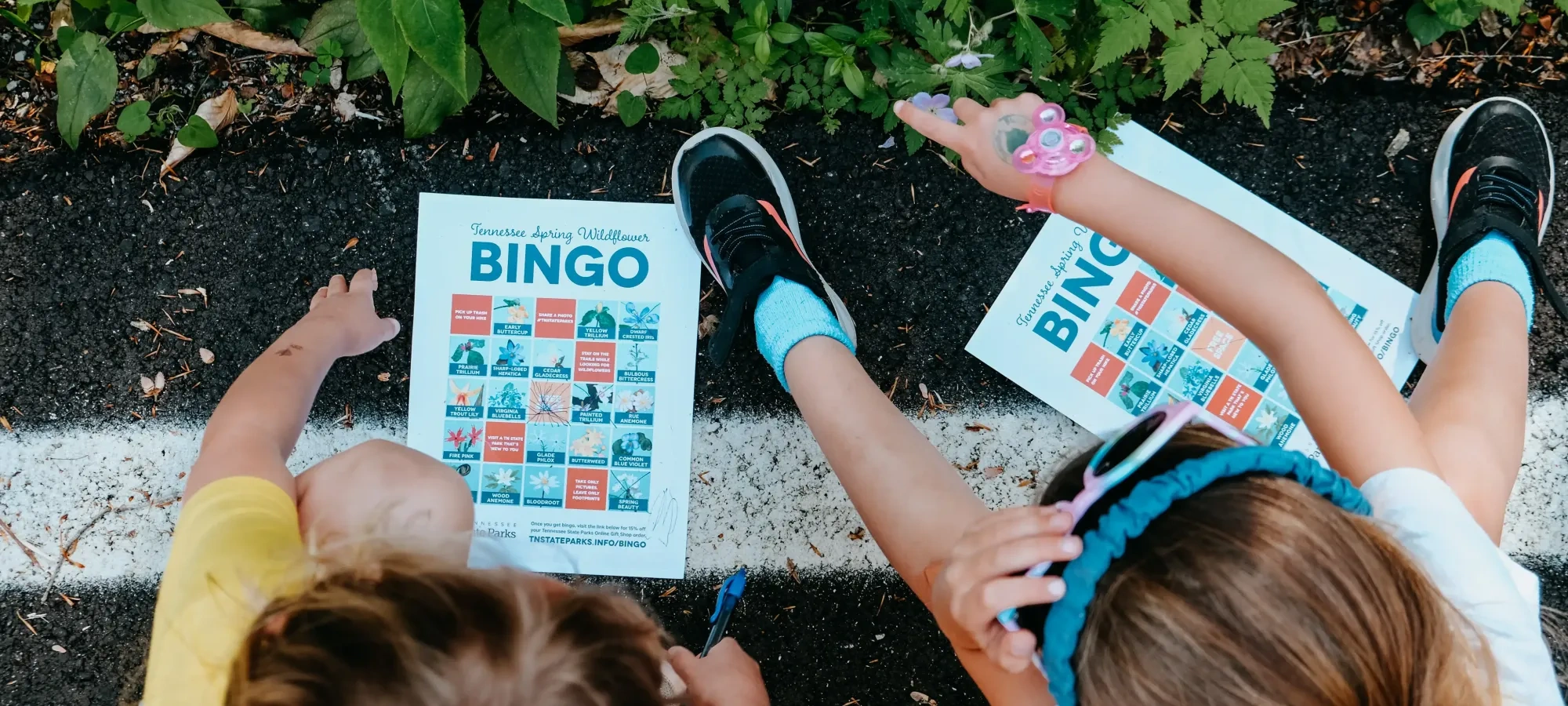 Two children seated on the ground playing a nature-themed bingo game, with the bingo cards and surrounding greenery visible.