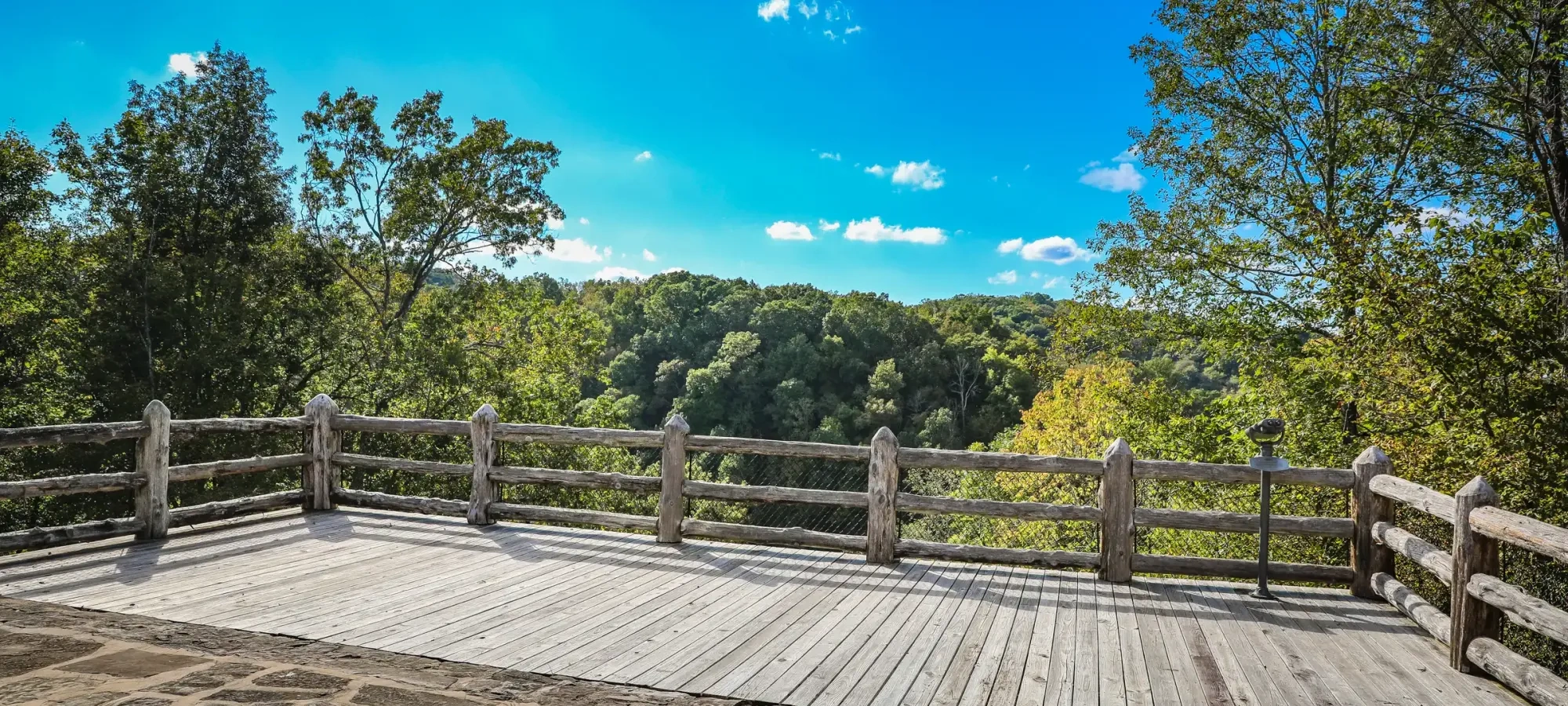 Wooden deck with rustic railing overlooking a lush forest under a bright blue sky.