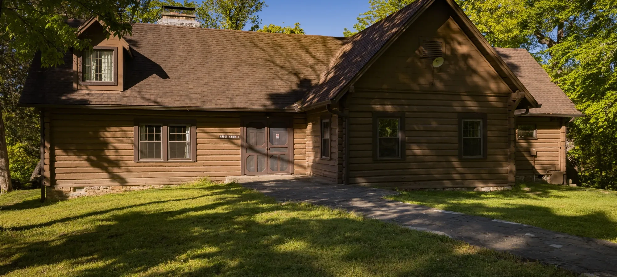 Brown log cabin with a sloped roof, surrounded by trees, under a clear blue sky.